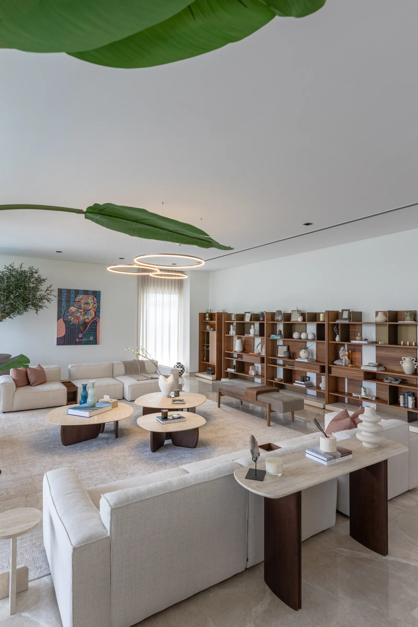Twyst Interiors.Dubai Hills GP open-plan living room with cream sofas, travertine tables, banana leaf overhead.