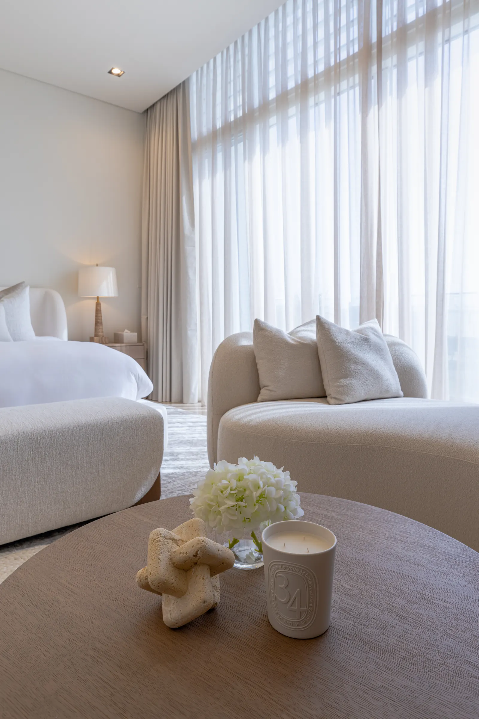Dubai Hills villa bedroom suite. Sheer curtain wall, circular boucle seating, low oak side table with hydrangeas.