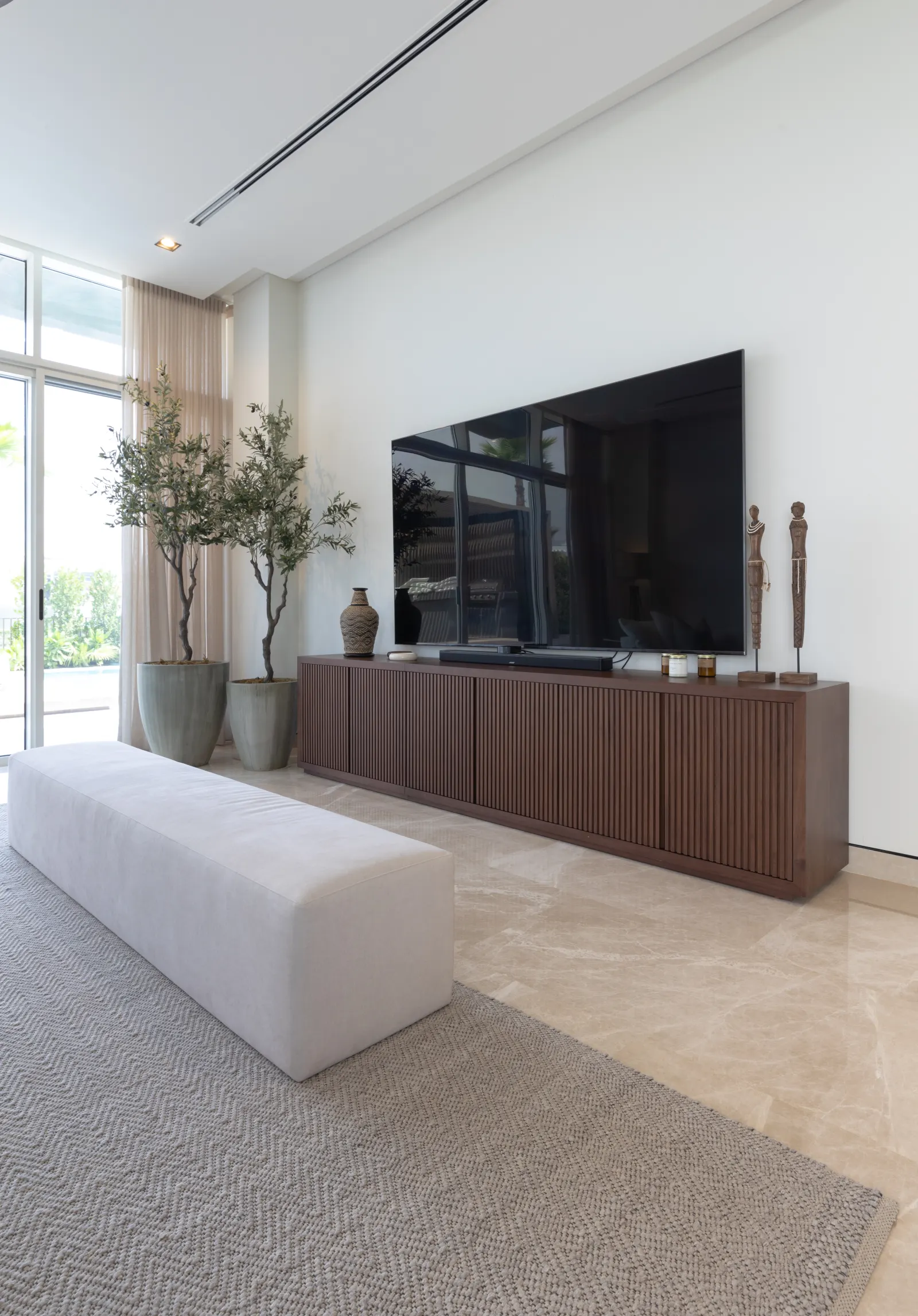 Dubai Hills villa living room. Double olive trees, walnut media console, boucle bench on textured rug, full-height curtain.