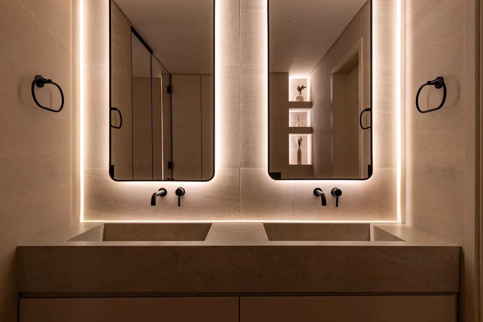 Twyst Interiors.Turk Villa primary bathroom with twin backlit mirrors, warm stone vanity, brushed black fittings.